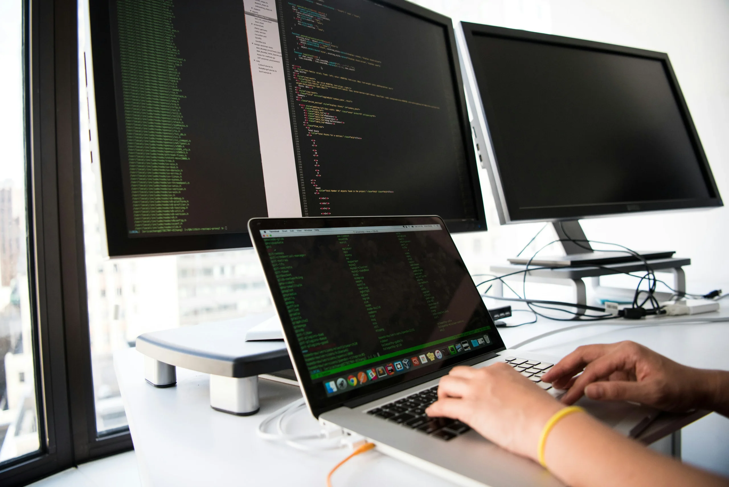 Vue rapprochée des mains d'une personne écrivant du code sur un ordinateur portable, connecté à deux grands écrans externes.