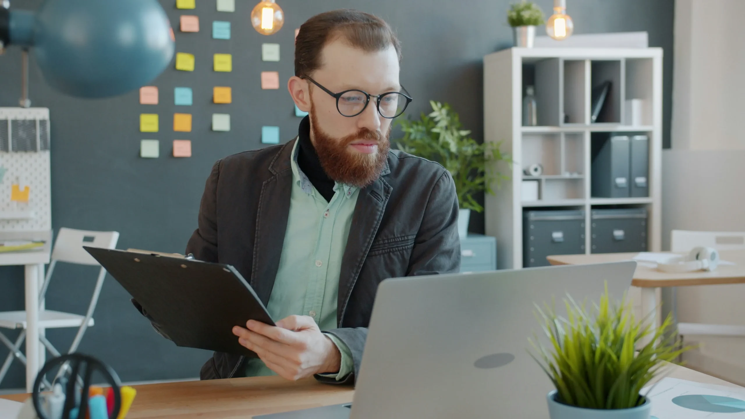Un homme barbu à lunettes assis à son bureau, regardant son ordinateur portable tout en tenant un porte-bloc avec des feuilles.