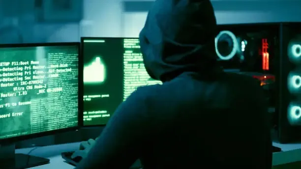Vue de dos d'un individu portant une capuche sombre, assis devant plusieurs écrans affichant des lignes de code.