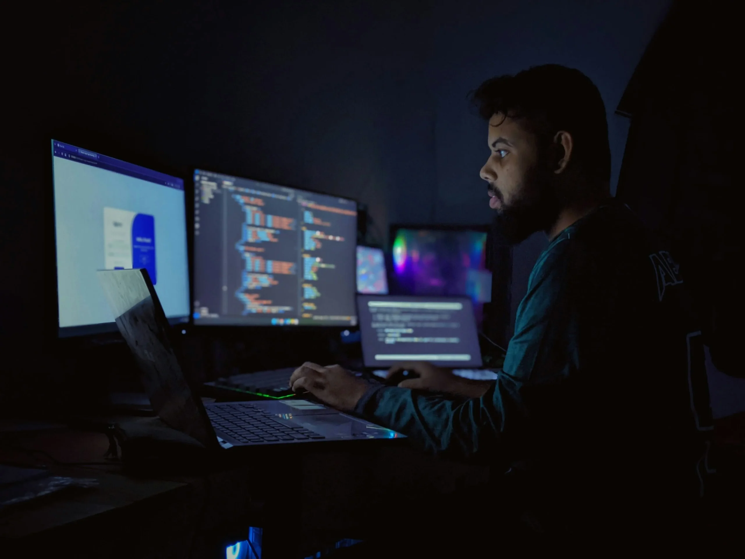 Un homme de profil travaillant de nuit dans une pièce sombre face à trois moniteurs et un ordinateur portable affichant du code.