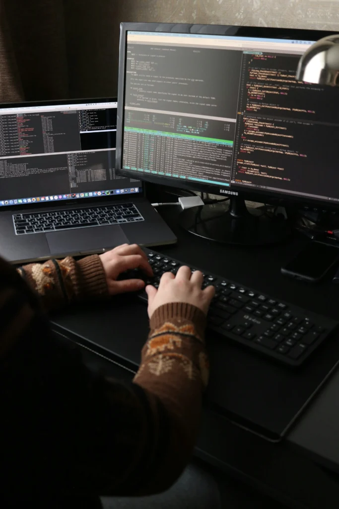 Vue d'une personne qui tape sur un clavier, installée à coté d'un ordinateur portable et devant un écran affichant des terminaux et du code.