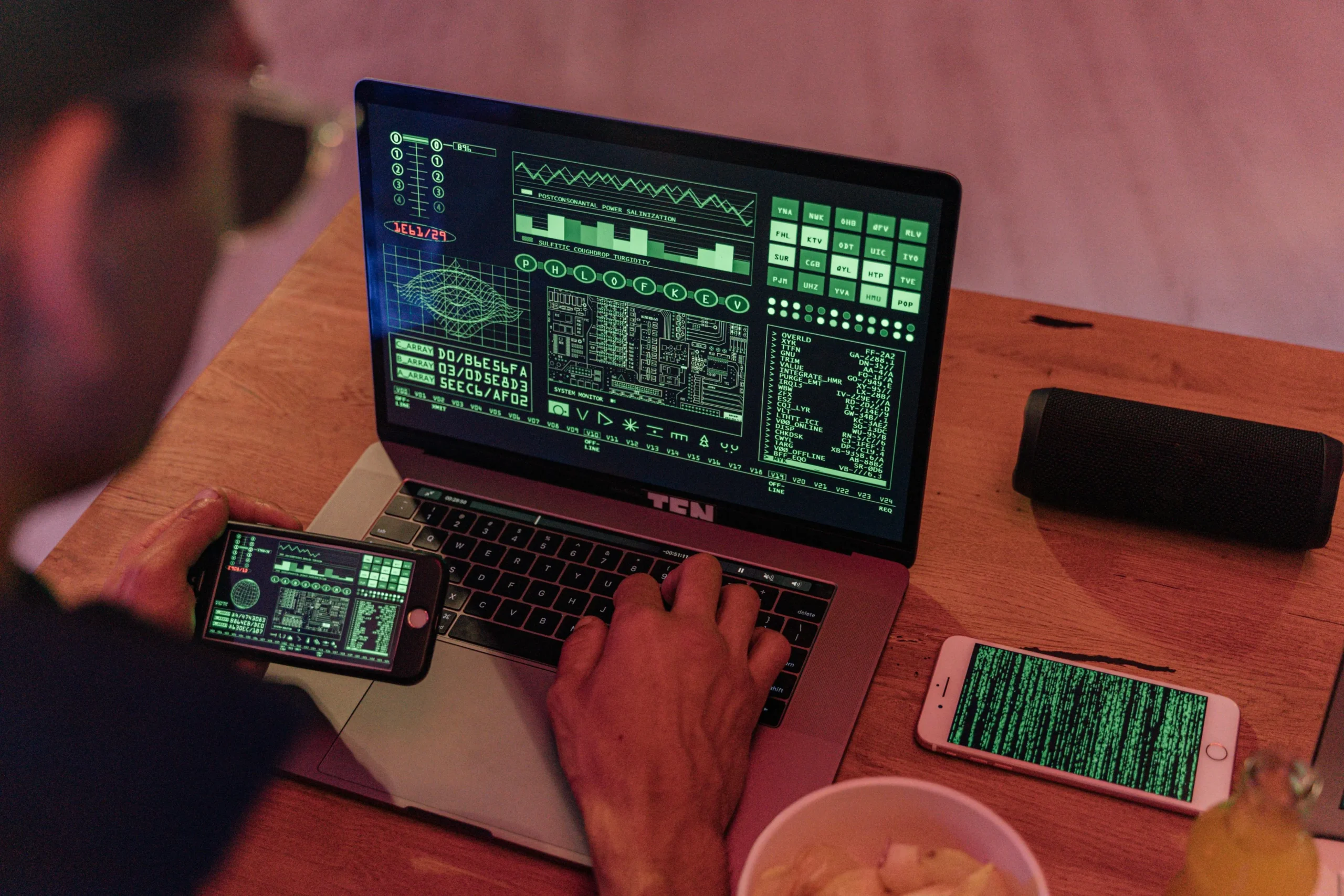 Vue en plongée d'une personne de dos utilisant un ordinateur portable, un smartphone à la main et un autre posé sur la table, tous affichant des lignes de code.