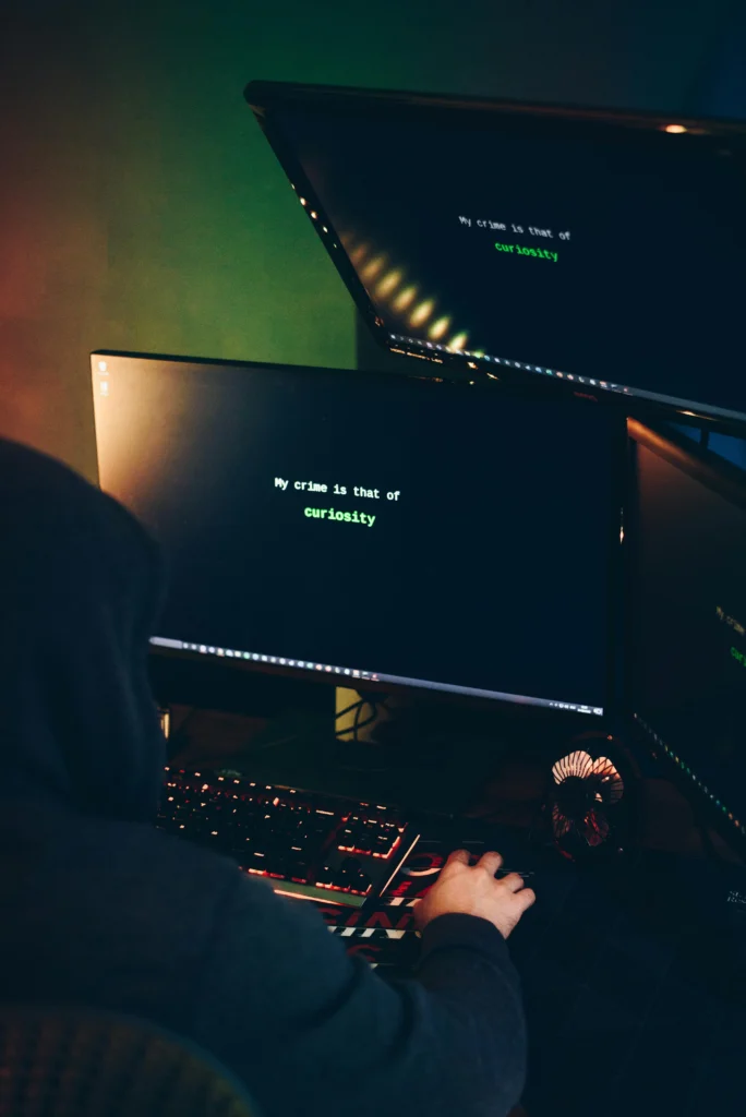 Vue arrière d'une personne portant une capuche noire, assise devant plusieurs écrans d'ordinateur affichant le texte vert « My crime is that of curiosity » sur fond noir.
