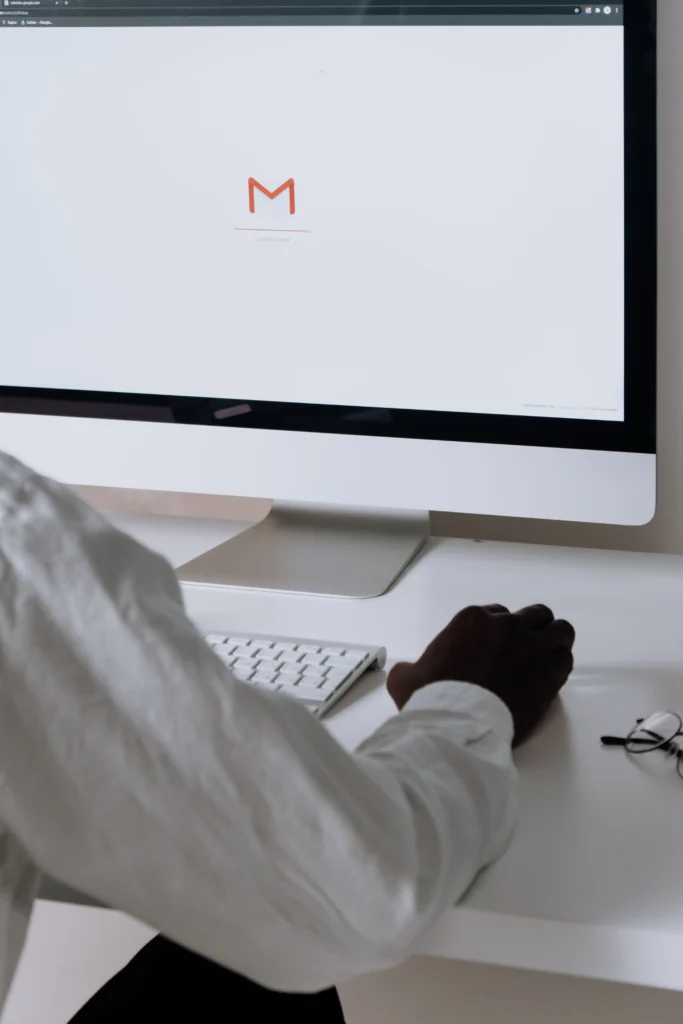 Vue de dos d'une personne, assise à un bureau et utilisant une souris. L'écran d'ordinateur devant elle affiche le logo de la messagerie Gmail.