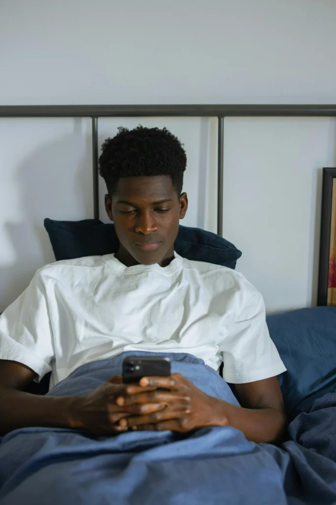 Un homme allongé dans son lit. Il regarde attentivement l'écran du smartphone qu'il tient à deux mains.