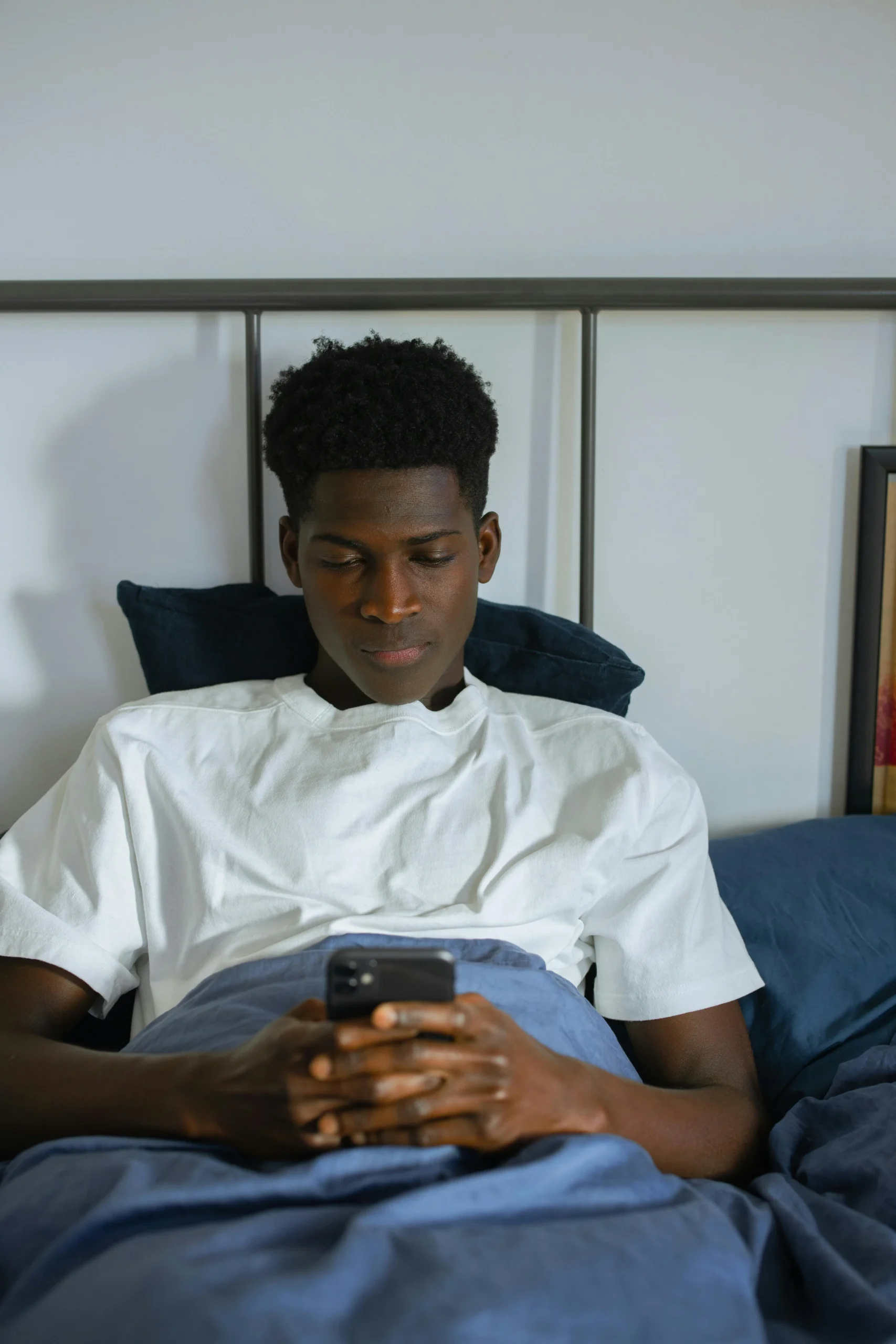 Un homme allongé dans son lit. Il regarde attentivement l'écran du smartphone qu'il tient à deux mains.