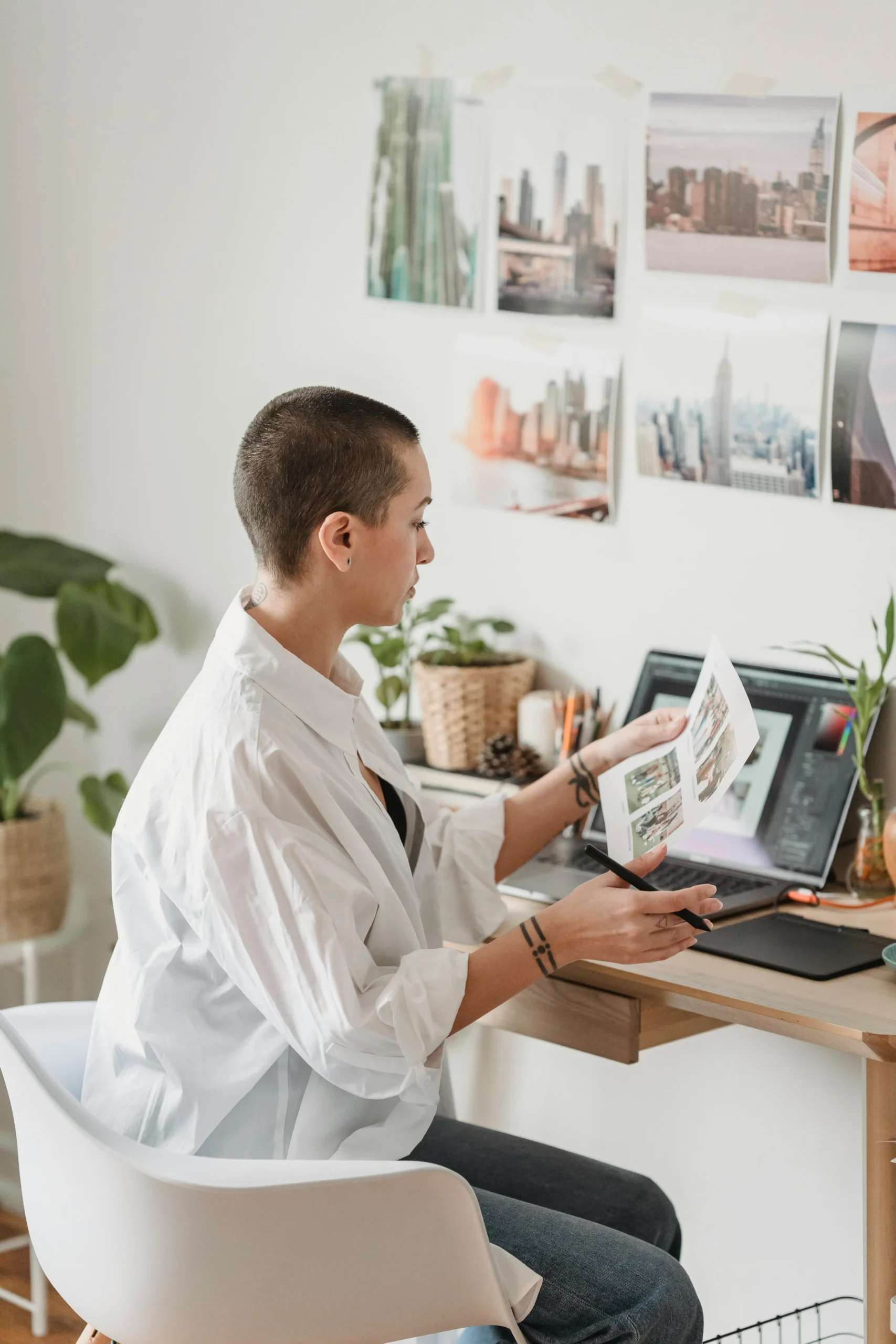 Une femme assise à son bureau de profil, examinant une feuille imprimée avec des photos devant un ordinateur portable ouvert.