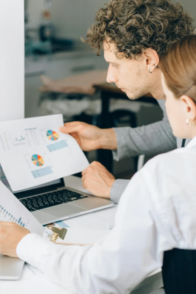 Un homme et une femme vus de profil à un bureau, analysant ensemble des graphiques imprimés sur papier à côté d'un ordinateur.