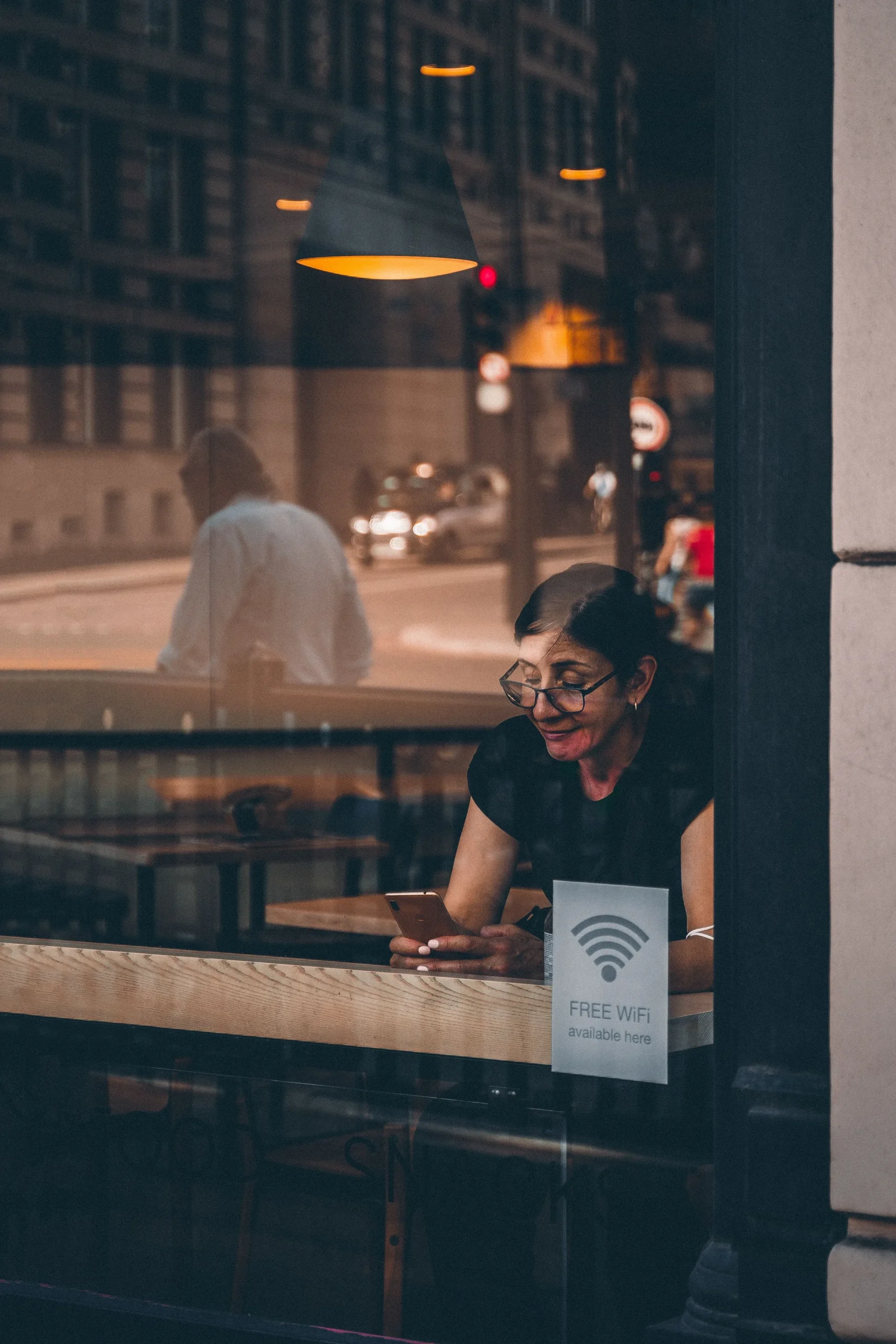 Une femme est assise à l'intérieur d'un café, regardant son smartphone. La scène est capturée de l'extérieur à travers une vitrine sur laquelle est collée une affichette indiquant « FREE WIFI available here ».
