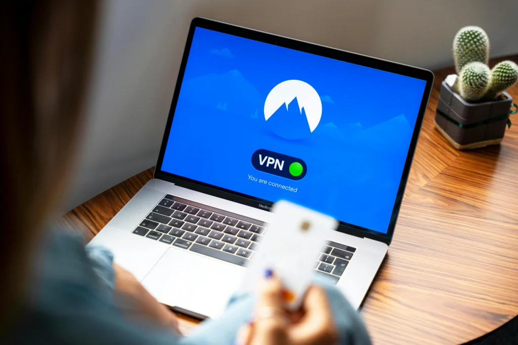 Vue par-dessus l'épaule d'une personne qui tient une carte bancaire face à un ordinateur sur une table. L'écran de l'ordinateur affiche une interface de connexion VPN active.