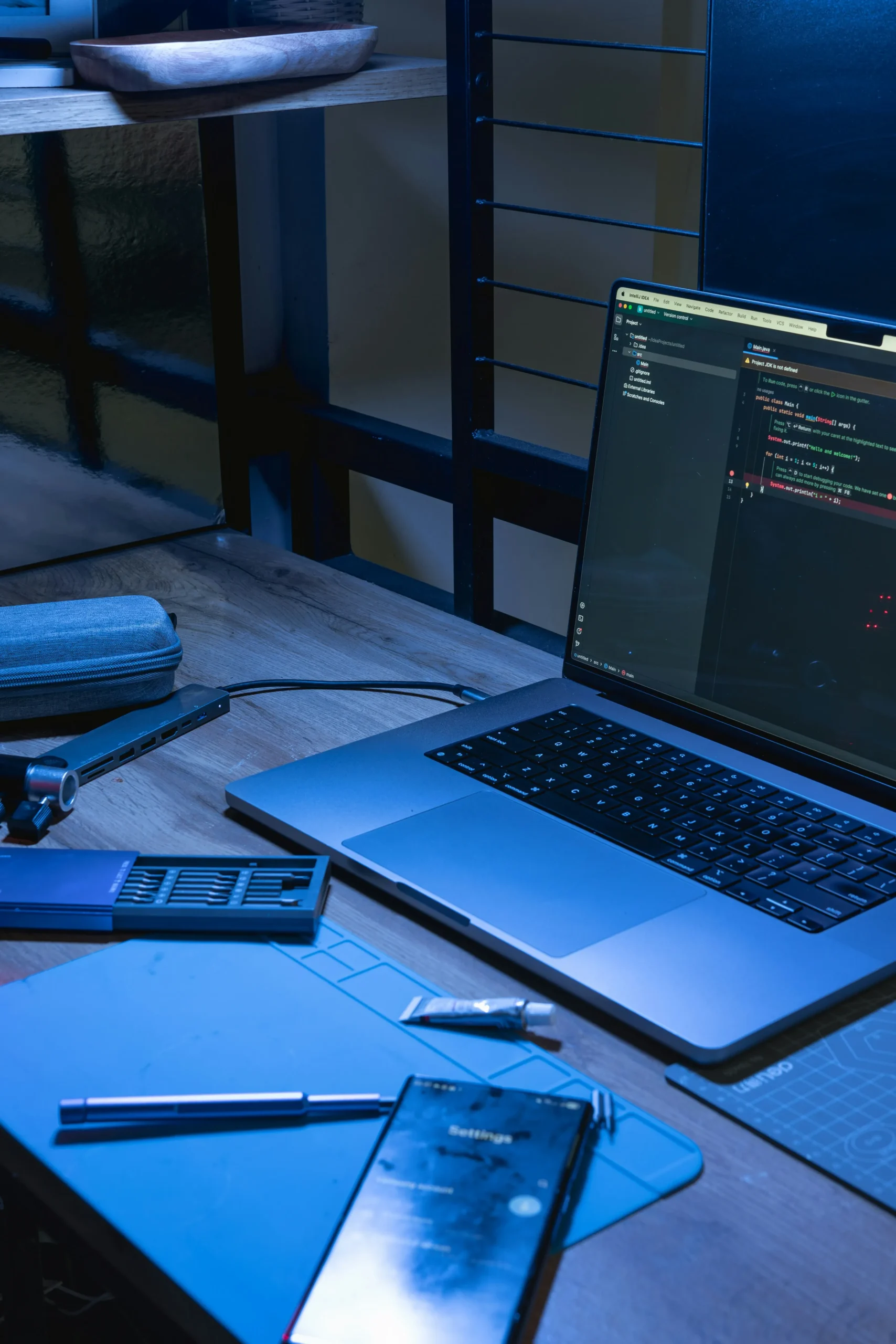 Un ordinateur portable ouvert affichant du code sur un bureau en bois éclairé par une lumière bleue, à côté d'un smartphone et d'outils.