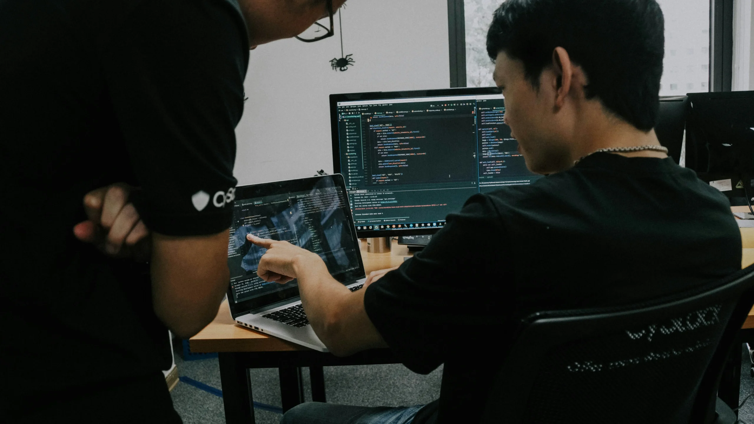 Deux personnes analysant et pointant du doigt des lignes de code sur un ordinateur portable et un écran fixe.